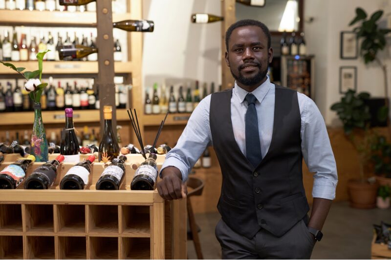 man standing in tasting room resting on a wine shelf comfortably dressed formally in a fancy shirt, vest and tie